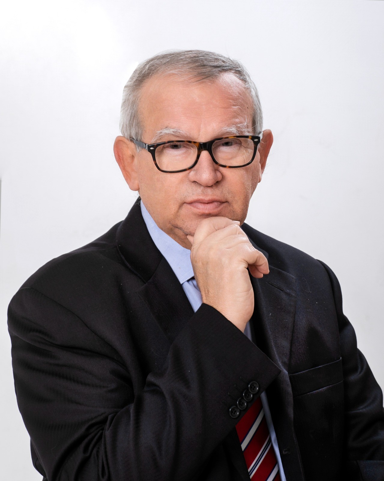 Luciano Bertolini in giacca scura, camicia azzurra e cravatta a righe rosse, occhiali tartarugati, mano sul mento