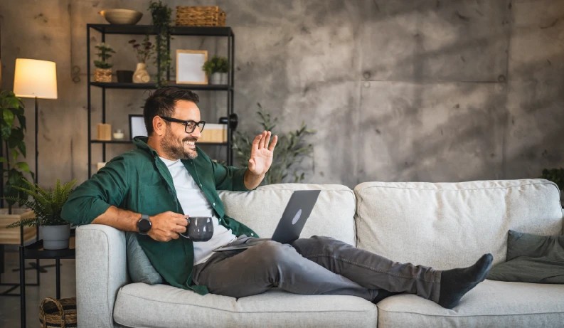 Uomo sorridente con occhiali e camicia verde saluta al portatile su un divano chiaro, tazza grigia in mano e muro di cemento